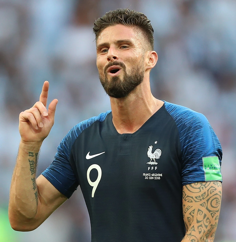 Olivier Giroud in a France national team match, wearing the number 9 shirt and gesturing with one finger raised.